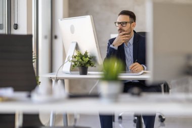 Takım elbiseli profesyonel bir işadamı modern ofis ortamında bilgisayar masasında özenle çalışıyor. Konsantrasyon ve verimlilik gösteriyor. Çevre parlak ve iyi düzenlenmiş..