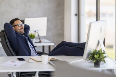 Takım elbiseli kendine güvenen bir işadamı modern bir ofiste ayakları masanın üzerinde rahatlar. Memnun ve memnun görünüyor, bir anlık başarı ya da işten uzaklaşma öneriyor..