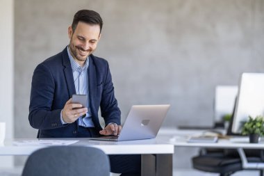 Takım elbiseli bir işadamı modern bir ofiste masa başında otururken akıllı telefonunu kontrol ederken gülümsüyor. Dijital aygıtlar arasında çoklu görev yapıyor.