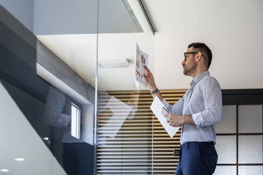 Bir iş adamı, odak ve stratejik planlamayı yansıtan çağdaş bir ofis ortamında cam bir duvarda sergilenen mali tabloları inceler..