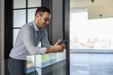 Modern ofis ortamında kendine güvenen bir işadamı, akıllı telefonunu kullanmaya dalmış. Görüntü profesyonelliği, iletişimi ve teknolojiyi parlak ve çağdaş bir çalışma alanında taşıyor..