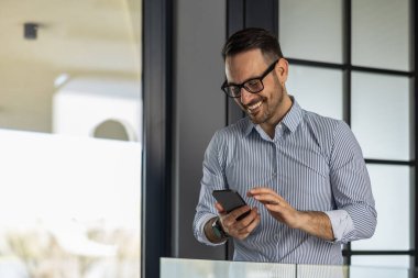 Gözlüklü ve çizgili tişörtlü neşeli bir işadamı gülümseyen bir akıllı telefon kullanıyor. Parlak, modern bir ofis ortamında duruyor. Pozitif ve üretken bir ruh halini yansıtıyor..