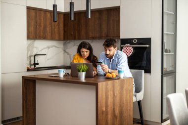 Genç bir çift mutfak adasında oturmuş, dikkatle dizüstü bilgisayara ve akıllı telefona bakıyor. Sahne, odaklanma ve takım çalışmasıyla modern bir mutfak ortamı yakalıyor..