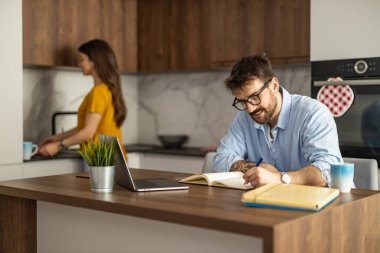Modern bir mutfakta dizüstü bilgisayar kullanırken notlar yazan bir adam. Bir kadın arka planda bir içki hazırlıyor, ev ofisi atmosferi yaratıyor..