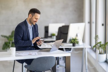 Takım elbiseli profesyonel bir adam, modern ve parlak bir ofis ortamında dizüstü bilgisayar üzerinde çalışırken akıllı telefonunu kullanıyor. Ofiste parlak mobilyalar ve bol miktarda doğal ışık bulunuyor..
