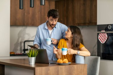 Bir çift, modern bir mutfakta birlikte sabah kahvesinin keyfini çıkarırken, dizüstü bilgisayar kullanırken sohbet ederken görülüyor. Ayar sıcaklık, bağlantı ve günlük rutin yayar..