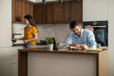 Neşeli bir çift modern bir mutfakta kaliteli zaman geçiriyor. Kadın yemek hazırlarken, erkek deftere sıcak ve rahat bir atmosfer yaratıyor..