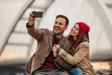 Mutlu çift kışın romantik bir açık hava anının tadını çıkarırken selfie çekiyor. Sıcak bir şekilde giyinmişler, birbirlerine kahkaha ve mutlulukla sarılıyorlar. İfadelerinde sevgi ve beraberlik gösteriyorlar..