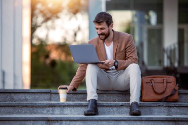 Genç bir profesyonel basamaklarda dizüstü bilgisayarla oturuyor, uzaktan gülümsüyor. Yanında bir kahve ve deri çanta var. Rahat ama üretken bir atmosfer yaratıyor..