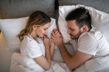 Huzur içinde uyuyan, yatakta el ele tutuşan genç bir çiftin huzurlu bir görüntüsü. Yumuşak ambiyans ve beyaz yatak rahatlama, samimiyet ve konfor sağlar..