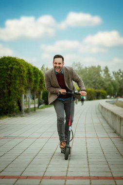 Neşeli bir adam mavi gökyüzünün altındaki bir park yolunda elektrikli scooter sürüyor. Sıradan bir spor ceket giyiyor ve görünüşe göre rahat bir sürüşten hoşlanıyor..