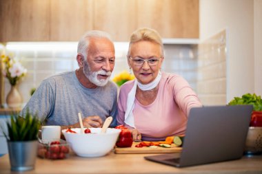 Mutlu son sınıf çifti, bir dizüstü bilgisayardaki online tarifi takip ederken modern mutfaklarında birlikte yemek pişirmeyi seviyor. Taze sebzeler ve çiçekler sıcak ve neşeli atmosferi güçlendirir..