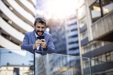 Takım elbiseli profesyonel bir işadamı cam korkuluklara yaslanıyor ve gülümseyen bir akıllı telefon kullanıyor. Modern mimari ile kentsel çevre kurumsal ve kendinden emin atmosferi geliştirir..