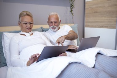 Üst düzey çift, modern teknoloji ve bağlantıyı sergileyerek, yatağında dizüstü bilgisayar ve tablet kullanıyor. Görüntü olgun arkadaşlığı, boş zamanı ve dijital çağı vurguluyor..