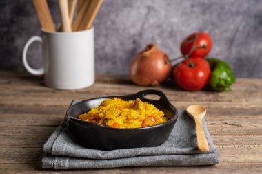 Köy usulü siyah tabak ve bir porsiyon etli paella. Arka planda sebzeler kullanılıyor..