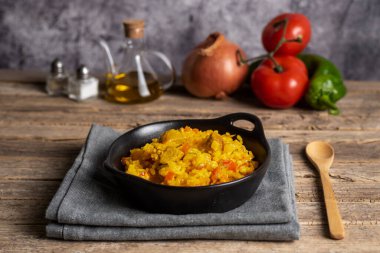 Köy usulü siyah tabak ve bir porsiyon etli paella. Arka planda sebzeler kullanılıyor..