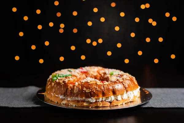 Roscon de reyes or king cake on a dark table and christmas lights in the background.