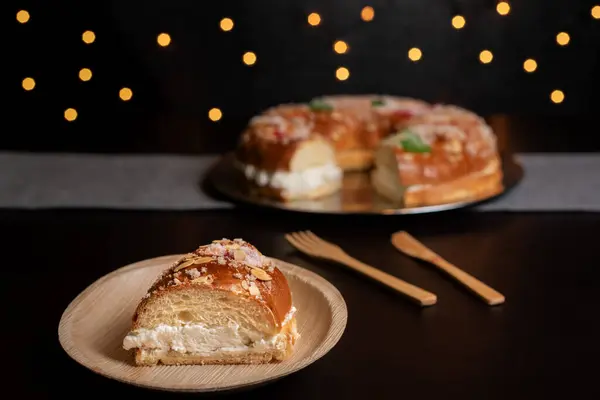 stock image Piece of roscon de reyes on a white and pink plate on a dark table with wooden cutlery, in the background a roscn de reyes and a cup of coffee out of focus and bokeh lights.