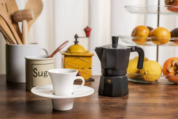 Cup of coffee on a kitchen table with a fruit bowl, a jug with wooden utensils, a coffee grinder and an Italian coffee maker.