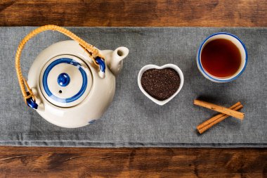 Porcelain teapot next to a small heart-shaped bowl with ground tea, a cup with tea and cinnamon sticks.