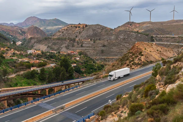 Truck with a refrigerated semi-trailer driving on a highway and a landscape with bridges, mountains, towns and wind turbines.