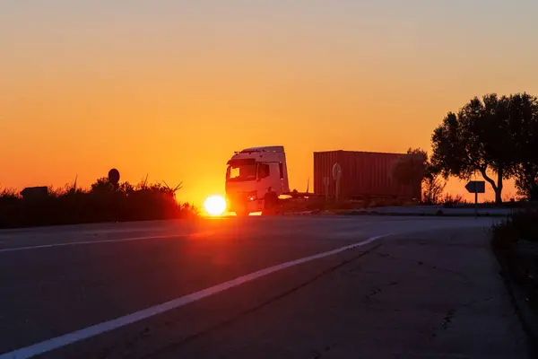 Truck with a semi-trailer with a maritime container driving on a conventional road with the sun very low.
