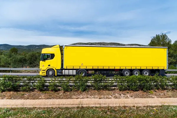 Tauliner yarı karavanlı bir kamyon tarlalar arasında bir otoyolda sürüyor, tamamen sarı..