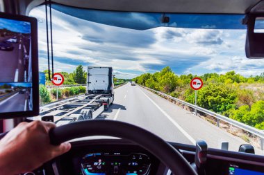 Karayolu üzerindeki bir kamyon dolusu trafik işaretinin sürücü koltuğundan kamyonların geçmesini yasaklarken, yasağı aşan bir başka kamyonun görüntüsü.