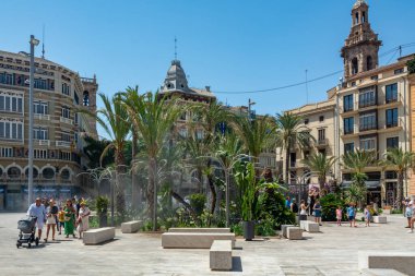 Valencia, İspanya - 12 Ağustos 2022: Turistler kalabalık bir şehir meydanında palmiye ağaçları ve tarihi cephelerin çerçevelediği çağdaş bir sis bahçesinin önünden geçtiler