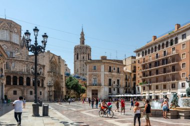 Valencia, İspanya - 12 Ağustos 2022: Turia Çeşmesi, pembe bazilika ve Gotik katedral cephesi tarafından çerçevelenmiş Plaza de la Virgen 'de gezen ve bisiklet süren ziyaretçiler.