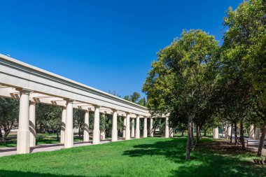 Valencia, İspanya - 12 Ağustos 2022: Beyaz sütun yolu kemerleri açık mavi gökyüzünün altında ağaçlarla ve manikürlü çimlerle kaplı yeşil bir şehir parkı çiziyor.