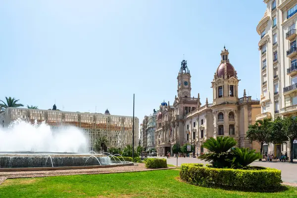 Valencia, İspanya - 12 Ağustos 2022: Süslü macenta kaplı belediye binası, hareketli Plaza del Ayuntamiento Meydanı 'ndaki dairesel bir fıskiyenin çağlayan su fıskiyeleri tarafından çerçevelenmiştir..