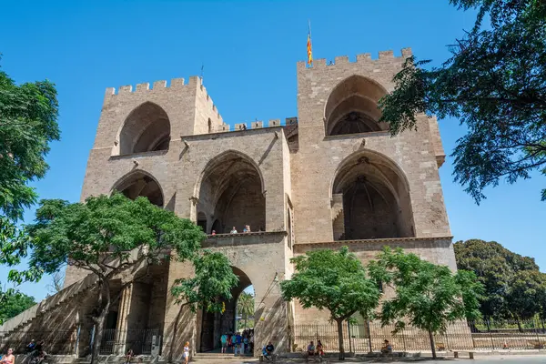 Valencia, İspanya - 12 Ağustos 2022: Ortaçağ Serranos Kuleleri 'nin arka kapısı yemyeşil sokak ağaçlarının üzerinde yükseliyor ve taş kemerlerinin altında yürüyen ziyaretçiler.