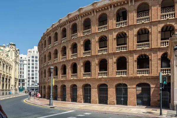 Valencia, İspanya - 12 Ağustos 2022: Plaza de Toros 'un güneşli tuğla kemerleri cadde boyunca ilerlerken ziyaretçiler açık mavi bir gökyüzü altında kapıların önünden geçiyorlar..