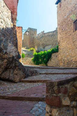 Güneş ışığı, berrak mavi gökyüzü tarafından çerçevelenmiş, crenelleştirilmiş kale duvarına kadar uzanan antik taş evlerle dolu bir ara sokağa girdi..