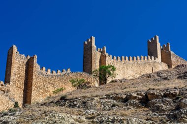 Tarihsel ortaçağ taş duvarları yüzyıllık tahkimatları sergileyen açık mavi bir yaz gökyüzünün altında engebeli bir yamaç boyunca uzanır..