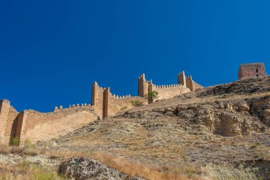 Tarihsel ortaçağ taş duvarları yüzyıllık tahkimatları sergileyen açık mavi bir yaz gökyüzünün altında engebeli bir yamaç boyunca uzanır..