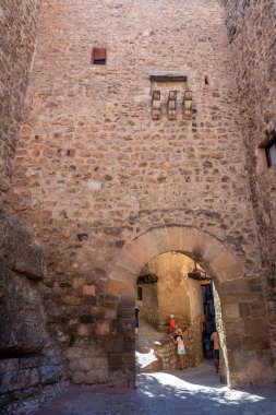 Albarakin, İspanya - 15 Ağustos 2022: Ziyaretçiler, engebeli uçurumlara yuva yapmış tarihi tepe kentinin dar kaldırımlı sokaklarına girerek, güneşli bir ortaçağ taş kemerinden geçiyorlar..