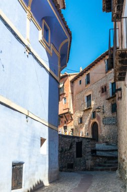 Albarakin, İspanya - 15 Ağustos 2022: Pastel mavi cepheli ve kırsal taş binalı günışığı ortaçağ sokağı, balkonlar ve kemerli pencerelerle çevrili dar kaldırım taşı yolu.