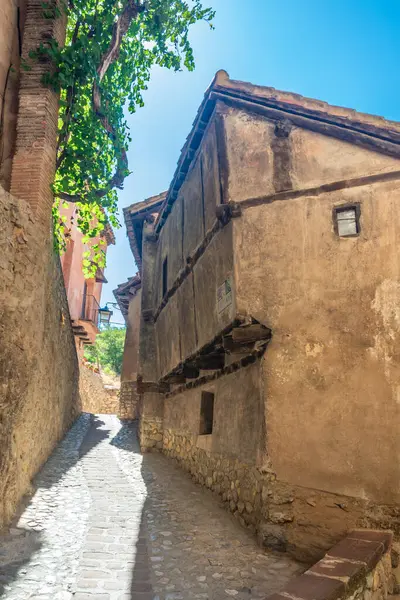 Dar bir kaldırım taşı geçidi, sarmaşıklara sarılı ahşap çerçeveli bir evin yanından geçiyor. Güneş ışığı, antik taş patikaya keskin gölgeler düşürüyor..