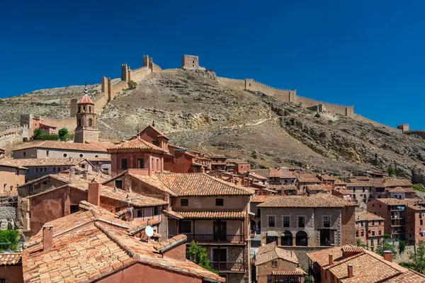 Terracotta çatıları eski kale duvarlarına doğru yükselir ve berrak mavi bir gökyüzünün altındaki kayalık yamacın tepesine tünemiştir..