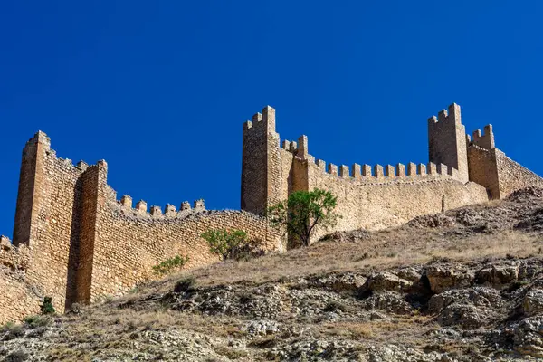Tarihsel ortaçağ taş duvarları yüzyıllık tahkimatları sergileyen açık mavi bir yaz gökyüzünün altında engebeli bir yamaç boyunca uzanır..