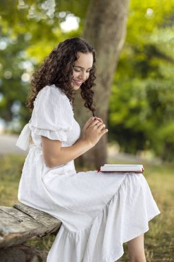 Yazın bir ağacın altındaki çayırda, ahşap bir bankta oturan bir kadın. Beyaz elbiseli genç kız tatilde doğada ders çalışıyor ve kitap okuyor.
