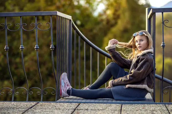 Bahçede kahverengi deri ceketli güzel bir kızın portresi. Gün batımında güneş gözlüklü genç kız parkta.