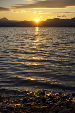 Gölün üstündeki dağların üzerinde güzel bir günbatımı. Ağaçlı bir deniz koyundaki suya yansıyan güneş ışınları.