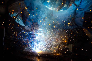 Kaynak maskeli, koruyucu giysili ve eldivenli bir adam atölyede çelik kaynaklıyor. Kıvılcımlı kaynak ateşi. Tehlikeli bir iş.