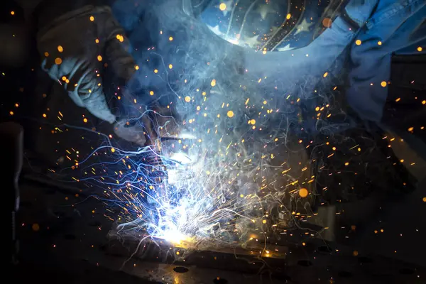 Kaynak maskeli, koruyucu giysili ve eldivenli bir adam atölyede çelik kaynaklıyor. Kıvılcımlı kaynak ateşi. Tehlikeli bir iş.