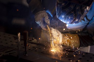 Kaynak maskeli, koruyucu giysili ve eldivenli bir adam atölyede çelik kaynaklıyor. Kıvılcımlı kaynak ateşi. Tehlikeli bir iş.