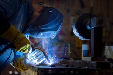Kaynak maskeli, koruyucu giysili ve eldivenli bir adam atölyede çelik kaynaklıyor. Kıvılcımlı kaynak ateşi. Tehlikeli bir iş.