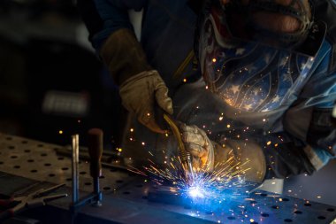 Kaynak maskeli, koruyucu giysili ve eldivenli bir adam atölyede çelik kaynaklıyor. Kıvılcımlı kaynak ateşi. Tehlikeli bir iş.